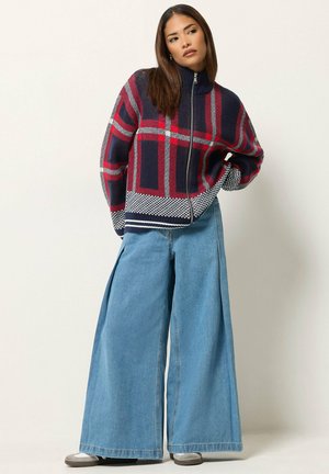 Femme portant un pull zippé à carreaux bleu marine et rouge et un jean large bleu, debout avec une main sur la hanche sur un fond uni.