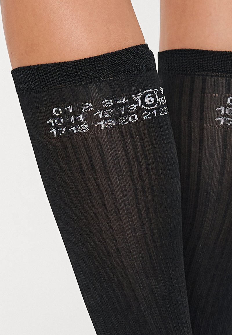 Chaussettes noires montantes avec une texture côtelée, ornées de motifs et de chiffres blancs le long de la bande supérieure.