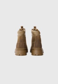 Botas tobilleras marrones con un upper de ante texturizado, un collar acolchado y suelas de goma gruesas con un patrón de tacos. Tablas de agarre en la parte trasera para facilitar su uso.