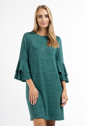 Frau mit glatten blonden Haaren, die ein grünes knielanges Kleid mit mehrlagigen Glockenärmeln trägt und vor einem schlichten weißen Hintergrund steht.