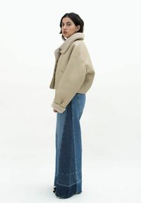 Korte beige leren jas met een pluizige beige kraag, gedragen met hooggekraagde blauwe denim flared broek met verticale panelen.