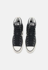 Zapatillas altas de canvas negras con suelas y costuras de goma blanca. Presentan un logo prominente, cordones planos y un collar acolchado para mayor comodidad.