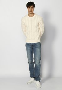 Suéter crema de punto trenzado con escote redondo, combinado con jeans de mezclilla azul. Modelo que lleva zapatillas blancas se encuentra de pie frente a un fondo liso.