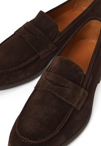 Mocasines de ante marrón oscuro con una tira para monedas, costuras visibles y interior de cuero marrón claro, presentados sobre un fondo blanco.