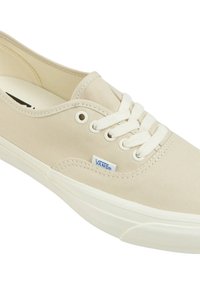 Sneaker in tela beige con lacci bianchi, caratterizzato da una suola curva, punta rotonda e un piccolo etichetta con logo blu sul lato.
