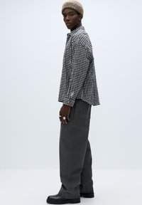 Grå ternet skjorte med lange ærmer og løs pasform, kombineret med mørkegrå vidde-bukser og sorte ankelstøvler. Modellen har en beige hue på.