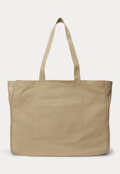 Μπεζ τσάντα ώμου από καμβά με δύο μακριές λαβές και ραμμένη ορθογώνια λεπτομέρεια στο μπροστινό μέρος, τοποθετημένη σε απλό λευκό φόντο.