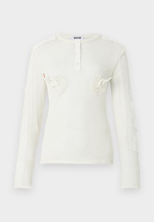 Modell kannab valget võrgust pika varrukaga pluusi valge rinnahoidja kohal, helesiniseid sirgete säärega pükse ja musti kõrgete kontsadega sandaale, seisab valgel taustal.