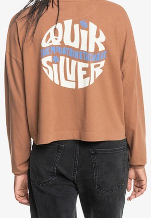 Personne portant un haut marron à manches longues court avec le texte blanc et bleu « Quiksilver The Mountain & The Wave » dans le dos et un jean noir.