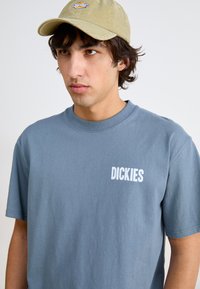 Pilkas medvilninis marškinėliai su trumpomis rankovėmis, kuriuose matomas baltas "DICKIES" logotipas. Dėvima su šviesiai rudaisiais šalmais, demonstruojant paprastą ir laisvalaikio dizainą.