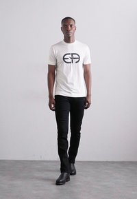Tricou alb cu logo negru, asociat cu pantaloni negri ajustați și cizme negre. Modelul stă în fața unui fundal neutru.