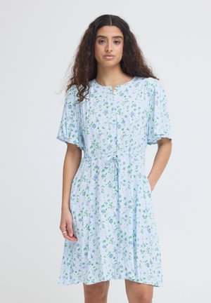 Hosszú, göndör hajú nő, világoskék, gombos, virágmintás ruhában, rövid ujjakkal és derékszalaggal, egyszínű háttér előtt állva.