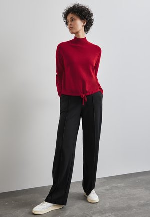 Roter Pullover mit langen Ärmeln, hohem Kragen und Kordelzugsaum, kombiniert mit schwarzen, weit geschnittenen Hosen und weißen Turnschuhen vor einem grauen Hintergrund.