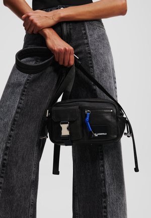 Black fabric crossbody bag with zippered compartments, silver hardware, and a blue cord accent. Paired with high-waisted grey jeans.