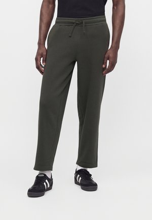 Homme portant un pantalon de survêtement vert foncé à cordon, des baskets noires et blanches, et un t-shirt noir, debout devant un fond uni.