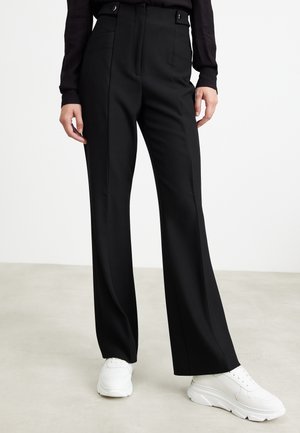 Person trägt schwarze, hoch taillierte, weit geschnittene Hose mit Knopf-Bundlaschen und weißen klobigen Sneakers vor neutralem Hintergrund.