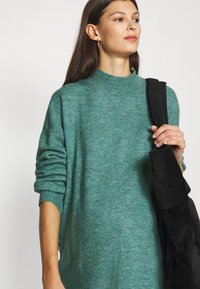 Pulover tricotat teal cu guler înalt cu nervuri și mâneci rulate, asociat cu o geantă de umăr neagră cu nervuri.