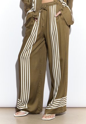 Pantaloni larghi verde oliva con righe bianche verticali e orizzontali, vita con coulisse e tasche, indossati con sandali bianchi a punta quadrata.