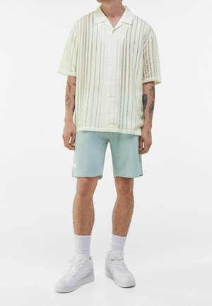 Camicia a maniche corte in tessuto traslucido giallo pallido con righe verticali, abbinata a pantaloni corti verde menta e sneaker bianche.