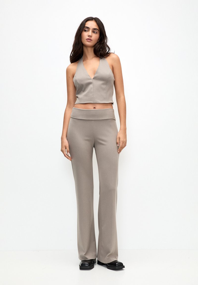 Beige verkürztes Neckholder-Top mit tiefem V-Ausschnitt, kombiniert mit hochtaillierten beigen Hosen. Der Stoff hat eine glatte Textur, und die Schuhe sind schwarz und klobig.