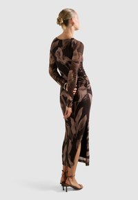 Donna con chignon biondo che indossa un vestito midi a maniche lunghe, marrone a fantasia, con spacco laterale e sandali con tacco a strappo, in piedi con le mani dietro la schiena.