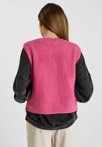 Fleece vest in helderroze, met een textuur oppervlakte, mouwloos ontwerp en ronde halslijn, gedragen over een donkere lange mouw shirt.