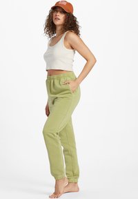 Lys grønne joggingbukser med elastisk talje og lommer, parret med en beige cropped tanktop og en brun kasket. Casual fit.