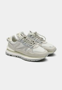 Zapatillas deportivas de color gris claro con una combinación de materiales lisos y texturizados, con una suela blanca gruesa y cordones reflectantes. Detalle sutil del logo en el lateral.