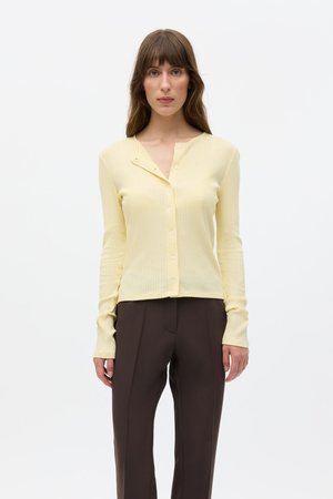 Žlutý žebrovaný kardigan s dlouhými rukávy a výstřihem do V, s knoflíkovou légou a krátkým lemem. Kombinován s tmavě hnědými kalhotami na míru.