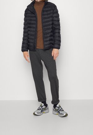 Osoba ubrana w czarną puchową kurtkę, brązową koszulę, ciemnoszare spodnie podwinięte przy kostkach, czarne skarpetki oraz srebrne buty do biegania z żółtymi i czarnymi detalami.