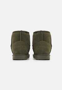 Botas de tobillo de ante verde con suela de goma plana y detalles de costura. Textura suave y forma redondeada visibles desde la vista trasera.