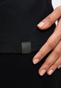 Χέρι με περιποιημένα νύχια που αγγίζει υφασμάτινη μαύρη επιφάνεια με μικρή ορθογώνια ετικέτα «BOSS» ραμμένη κοντά στην άκρη.