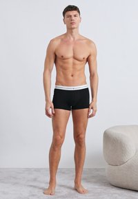 Black cotton boxer briefs with a white elastic waistband featuring a logo, fitted design, and smooth texture. Model poses upright.