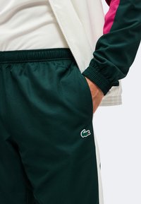 Άτομο με σκούρο πράσινο παντελόνι Lacoste με κεντημένο λογότυπο κροκοδείλου, λευκό και πράσινο μπουφάν, χέρι στην τσέπη του παντελονιού.