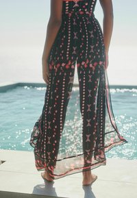 Zwarte doorzichtige maxi-jurk met een bloemenpatroon in rood en roze, met een cinched taille en zijsplitten, gecombineerd met blote voeten.