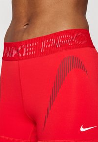 Crvene atletske kratke hlače s prozračnim elastičnim pojasom koji ima kontrastni bijeli "NIKE" logo i vertikalni uzorak prugastih linija sa strane.