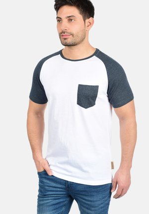 Hvid og navy raglan t-shirt lavet af bomuld, med en brystlomme, korte ærmer og en lige nederste kant. Blå denim jeans synlige.