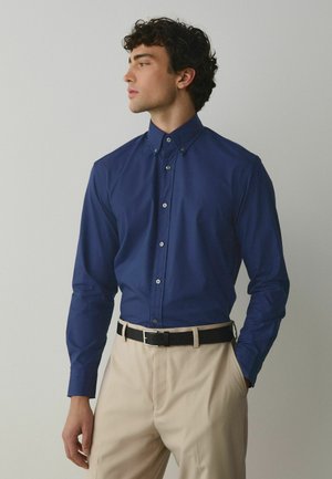 Man wearing a navy blue button-up shirt tucked into beige pants with a black belt, standing with one hand in pocket looking left.