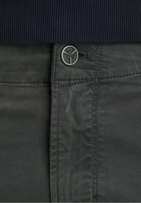 Detailní záběr na tmavě šedý pas kalhot s kovovým knoflíkem vyraženým s nápisem "PME Legend Jeans" a částí námořnického hřejivého svetru nahoře.