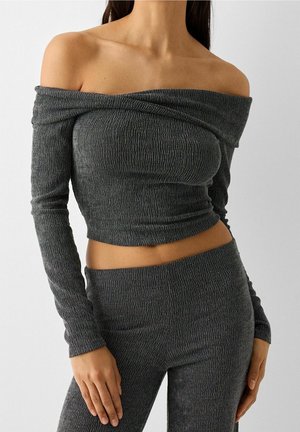 Frau trägt ein passendes, dunkelgraues, strukturiertes, schulterfreies Langarm-Crop-Top und hochtaillierte, eng anliegende Hosen vor neutralem Hintergrund.