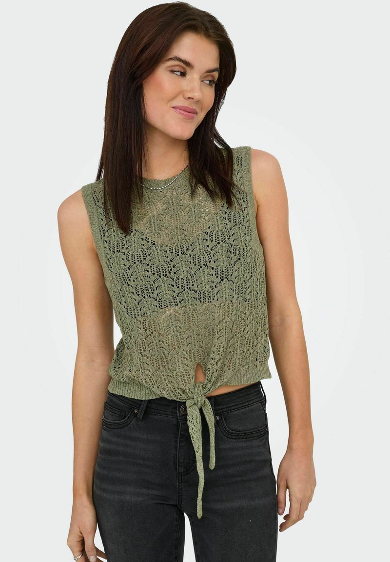 Top corto verde lavorato a pizzo con design senza maniche e dettaglio con fiocco frontale, abbinato a jeans neri. Motivo aperto e testurizzato su tutta la superficie.