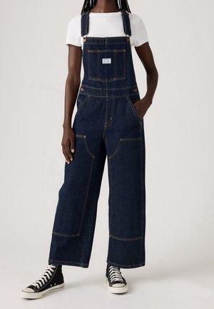 Femme portant une salopette en jean bleu foncé à jambes larges, un t-shirt blanc à manches courtes et des baskets montantes noires, main dans la poche.