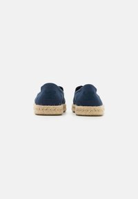 Mørkeblå kanvas slip-on sko med en afrundet bagdel, der har en sål af naturlig jute reb og præget logo-detail langs overdelen.