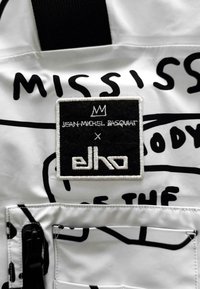 Fehér szövet fekete absztrakt mintákkal, amelyen egy téglalap alakú fekete folt látható fehér hímzéssel, rajta a "JEAN-MICHEL BASQUAT x elho." felirattal.