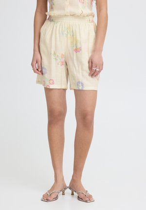Woman wearing cream elastic-waist shorts with pastel floral embroidery and snakeskin-patterned heeled sandals on a plain background.