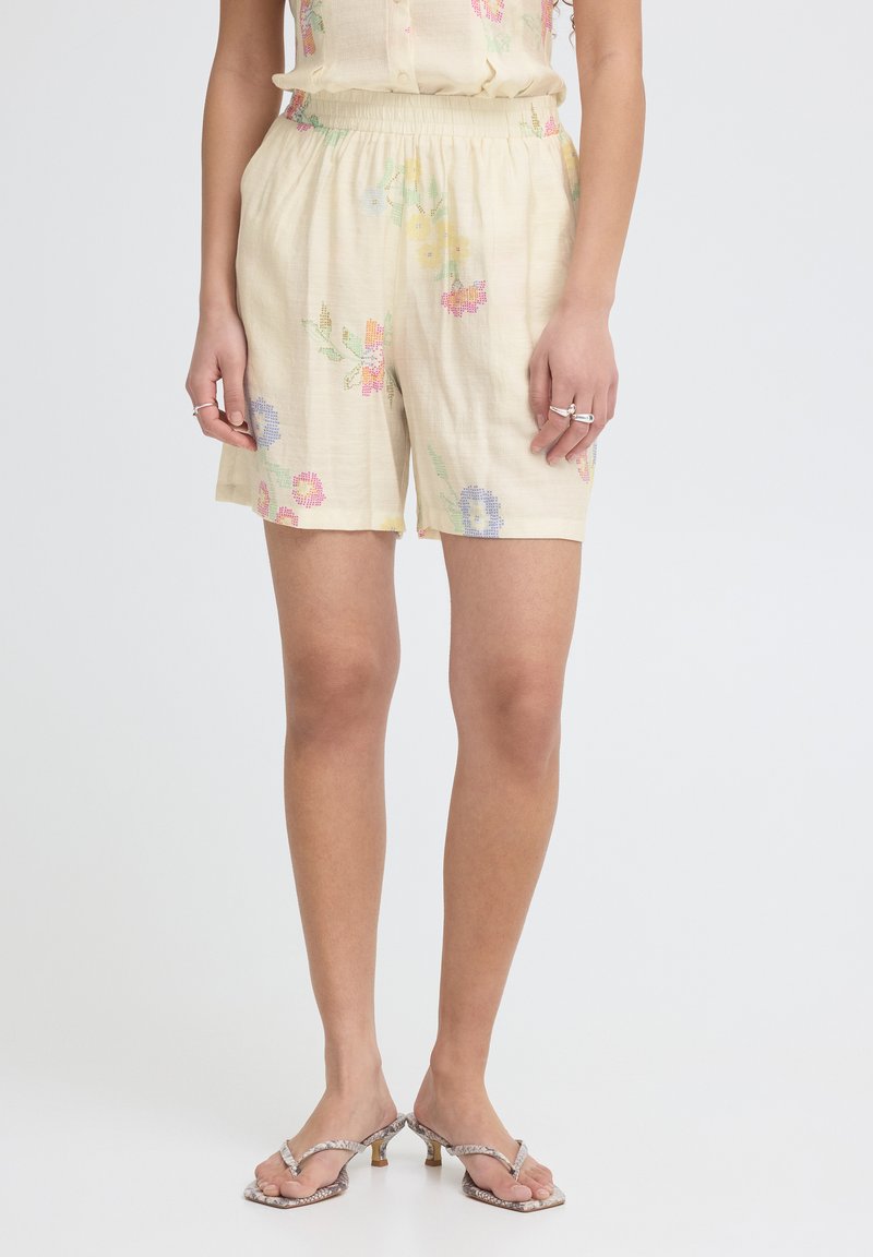 Femme portant un short crème à taille élastique avec une broderie florale pastel et des sandales à talons à motif peau de serpent sur un fond uni.