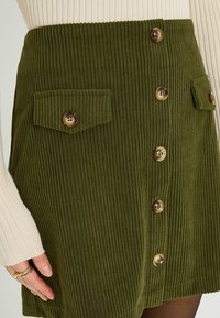 Jupe en velours côtelé verte avec une texture côtelée, un devant à boutons et deux poches poitrine avec des boutons marron.