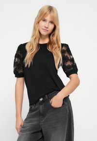 Μαύρο μπλουζάκι με διάφανα, κεντητά μαύρα φουσκωτά μανίκια με λουλούδια. Το μοντέλο φοράει γκρι ψηλόμεσα τζιν, με τα χέρια στις τσέπες.
