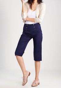 Marineblauwe cropped broek met knoopdetails aan de voorkant, gecombineerd met een aangesloten wit tanktopje en lichtgouden cardigan, afgewerkt met blushkleurige oxford schoenen.