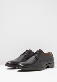 Chaussures habillées en cuir noir avec une finition lisse, bout rond, détail à lacets et un petit talon empilé. L'intérieur est doté d'un revêtement marron.